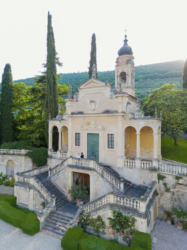 Ancient baroque chapel surrounded by lush greenery and cypress trees, emphasizing historical architecture and scenic beauty.