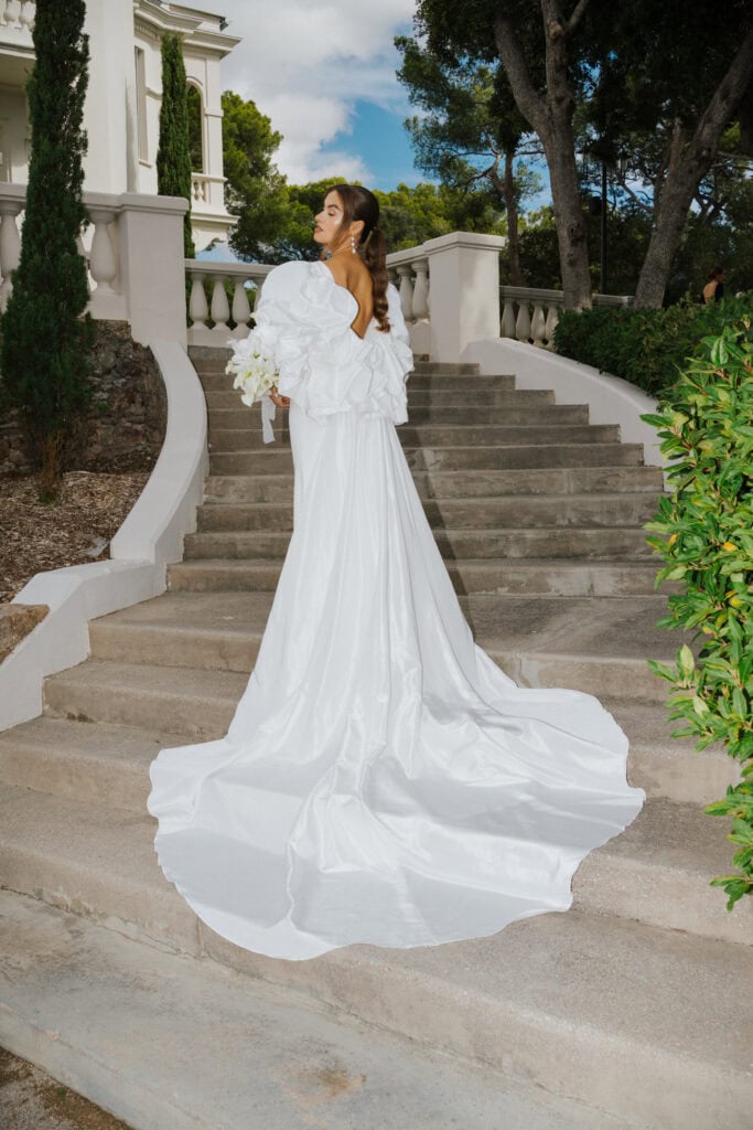 Elegantes weißes Hochzeitskleid mit Puffärmeln und einer langen Satinschleppe auf einer Steintreppe im Freien unter strahlend blauem Himmel.