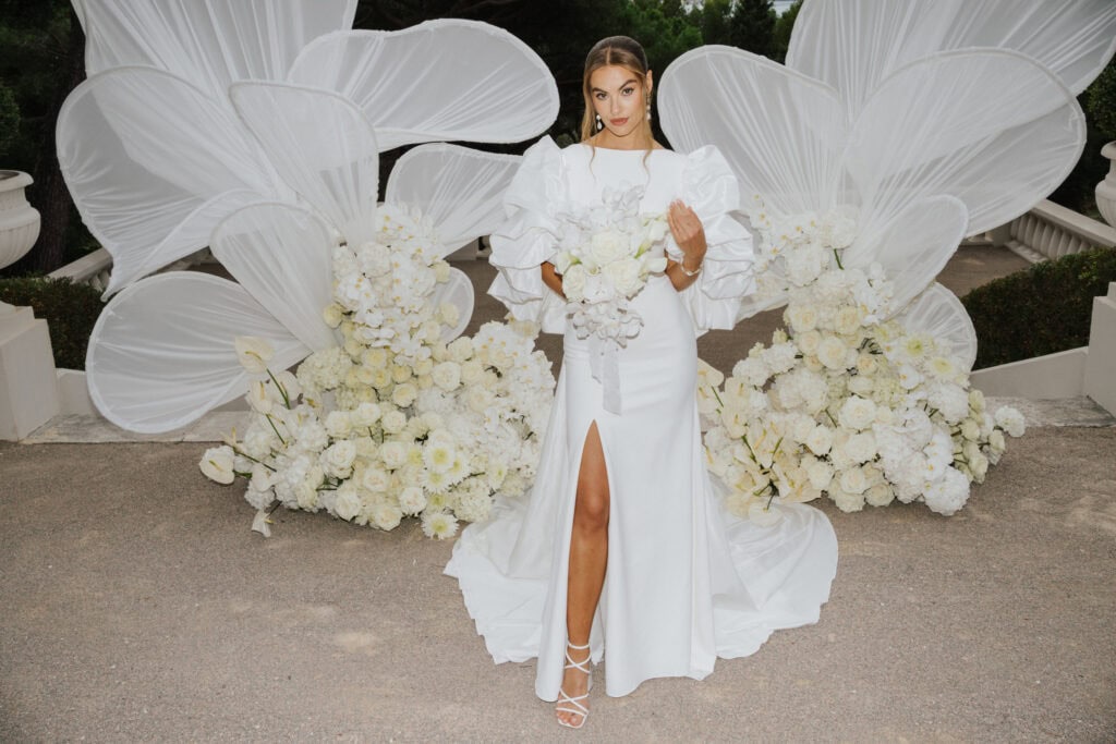 Elegantes weißes Hochzeitskleid mit dramatischen Puffärmeln und hohem Schlitz, gestylt mit Blumenbouquet, vor einem atemberaubenden Blumenhintergrund mit großen weißen schmetterlingsförmigen Akzenten.