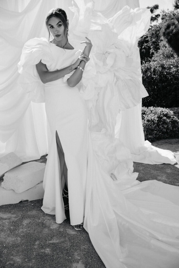 Elegante Frau in einem atemberaubenden weißen Kleid mit dramatischen Puffärmeln und einem hohen Schlitz, die im Freien vor einem Blumenhintergrund in Schwarz und Weiß posiert.