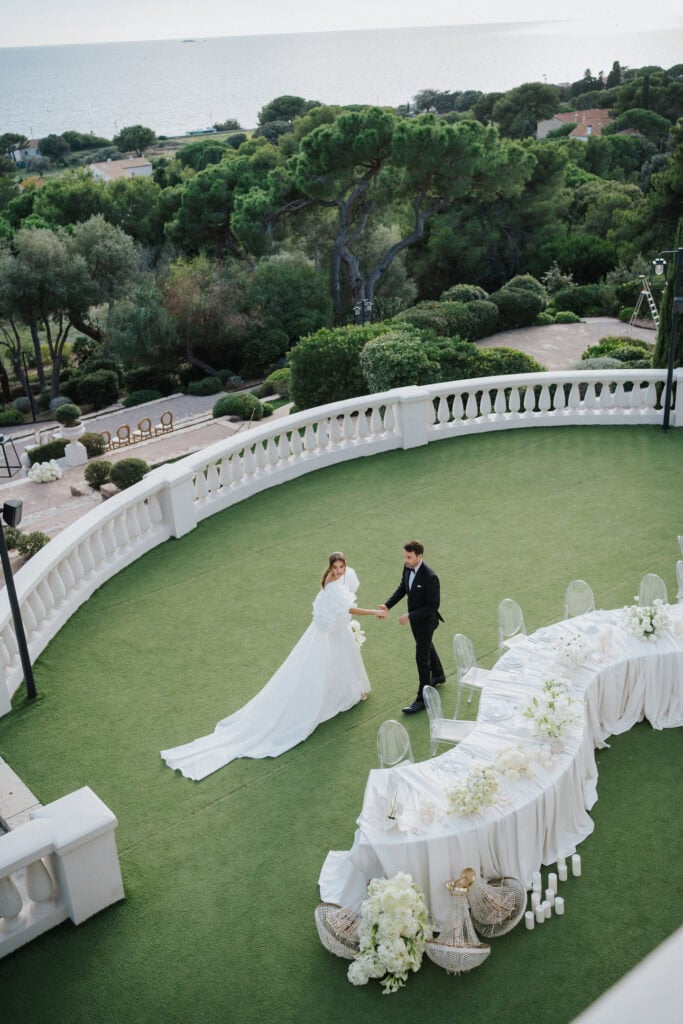 Eleganter Hochzeitsempfang im Freien auf üppig grünem Rasen mit Blick aufs Meer, weißer Blumendekoration und stilvollen transparenten Stühlen.