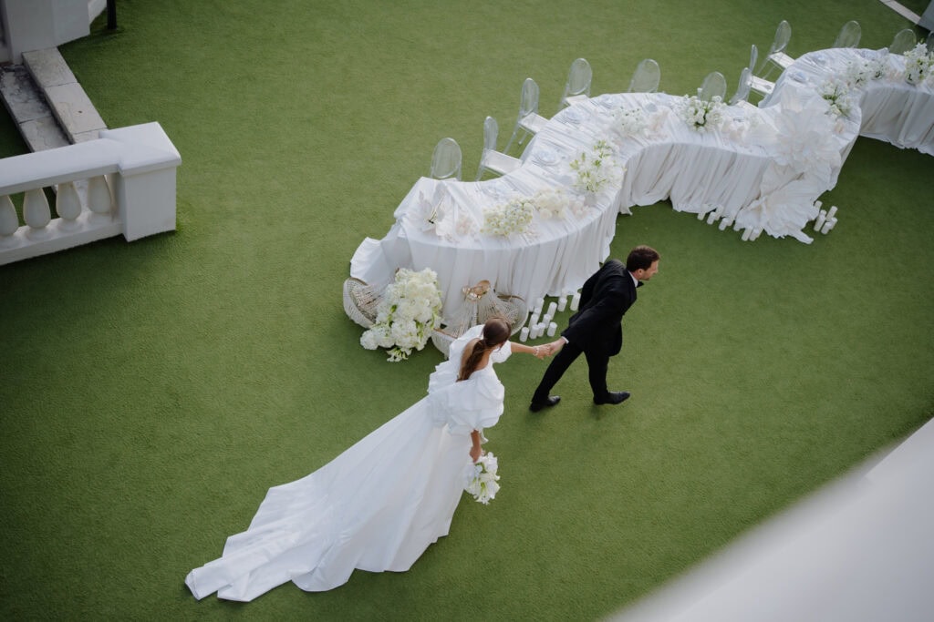 Eleganter Hochzeitsempfang mit einem langen weißen Tisch, der mit Blumenarrangements geschmückt ist, umgeben von durchsichtigen Geisterstühlen, auf einem üppigen grünen Rasen in Castel Bay.