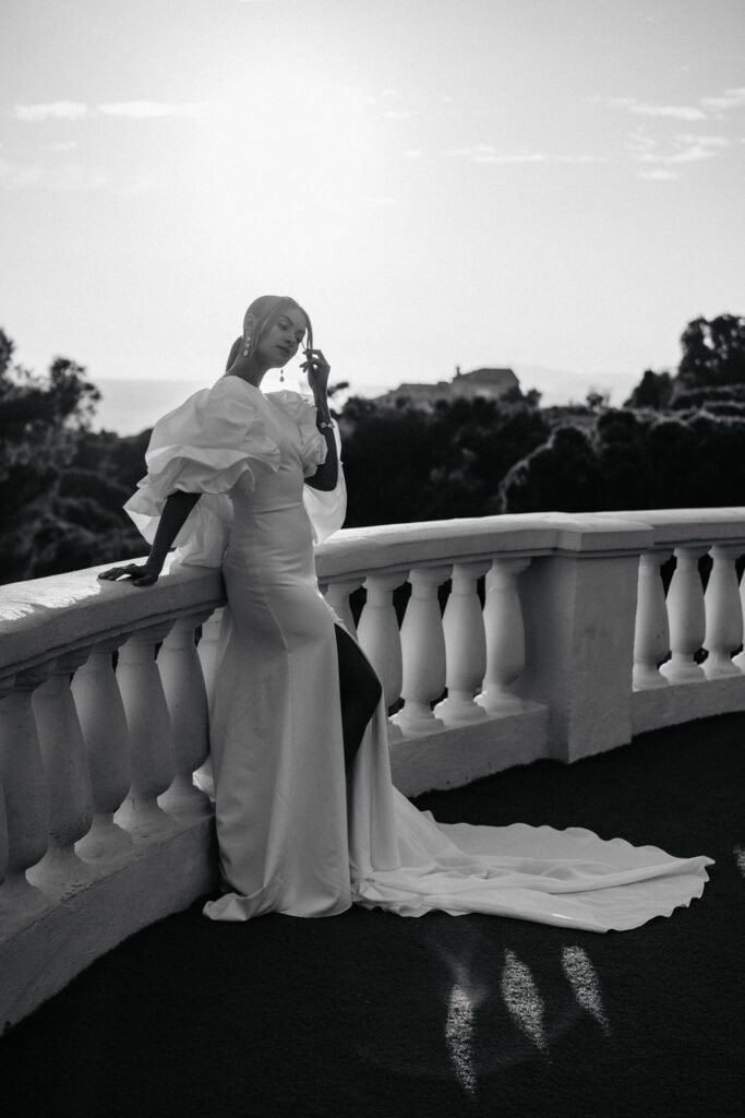Elegante Frau in einem atemberaubenden weißen Kleid posiert auf einem Balkon mit malerischer Landschaft Hintergrund, präsentiert luxuriöse Designer-Kleid und anspruchsvollen Stil.