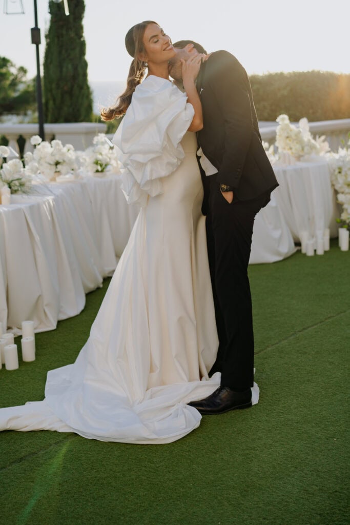 Ein freudiger Moment auf einem Hochzeitsempfang, bei dem sich Braut und Bräutigam bei Sonnenuntergang im Freien umarmen. Elegante weiße Blumenarrangements und Kerzen schaffen ein romantisches Ambiente in der Castel Bay.