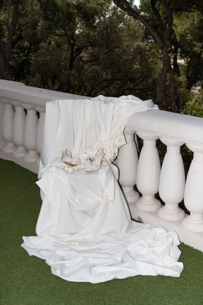 Elegantes weißes Hochzeitskleid und Accessoires, drapiert über ein weißes Balkongeländer mit üppigem Grün im Hintergrund.