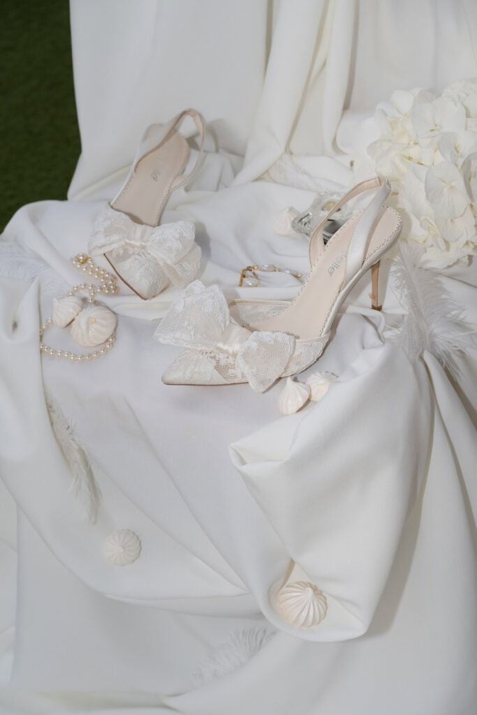 Elegante Brautschuhe mit Spitzenschleifen auf einem weißen Satintuch, umgeben von Hochzeitsaccessoires und Blumen.