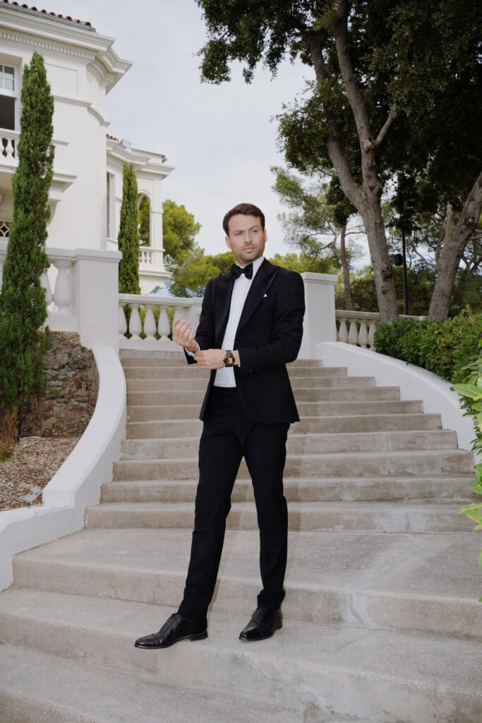Eleganter Bräutigam im schwarzen Smoking steht auf der Außentreppe eines luxuriösen Herrenhauses
