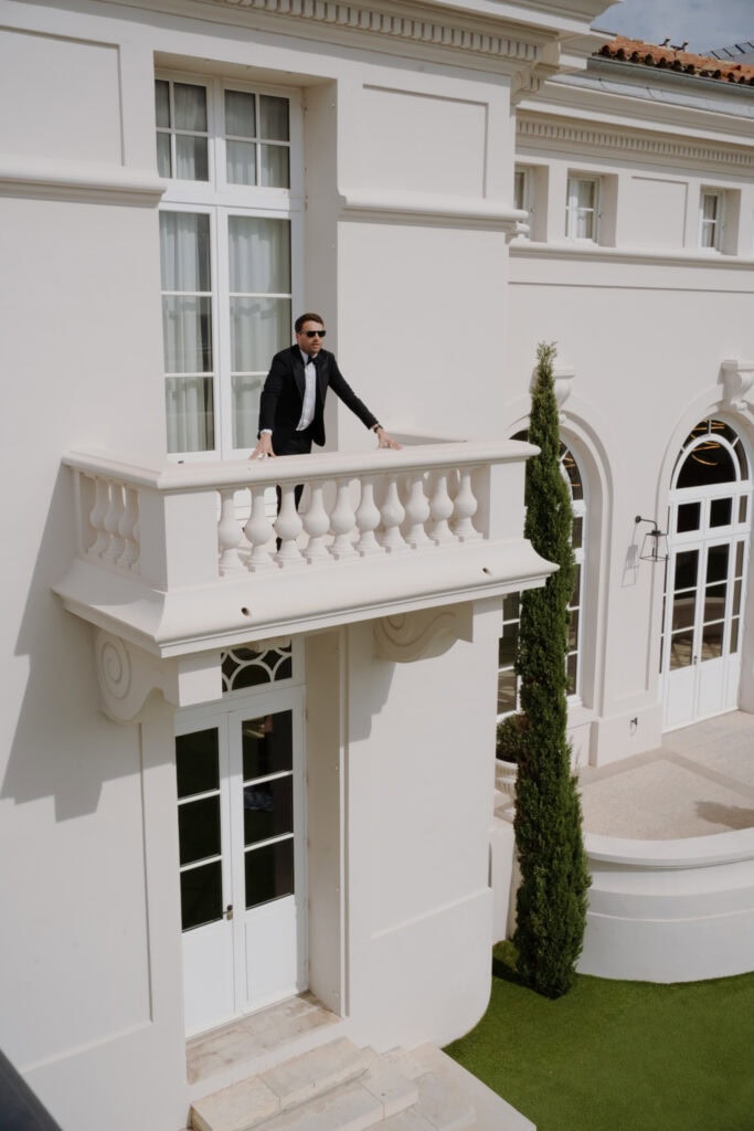 Stilvoller Bräutigam im schwarzen Anzug auf einem weißen Balkon einer eleganten Villa, der einen raffinierten und künstlerischen Moment bei Tageslicht einfängt.