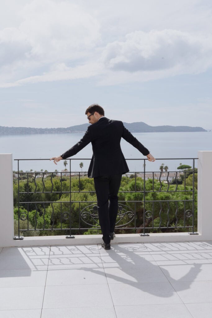 Ein stilvoller Bräutigam in einem maßgeschneiderten schwarzen Anzug genießt an einem teilweise bewölkten Tag den Blick vom Balkon auf einen ruhigen See und üppiges Grün.