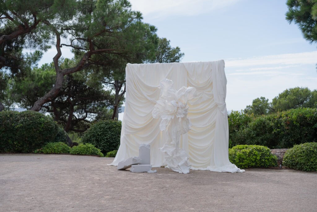 Eine moderne Hochzeitskulisse im Freien mit einem fließenden weißen Hintergrund und zerknitterten Skulpturen vor üppigem Grün und hohen Bäumen an einem klaren Tag.