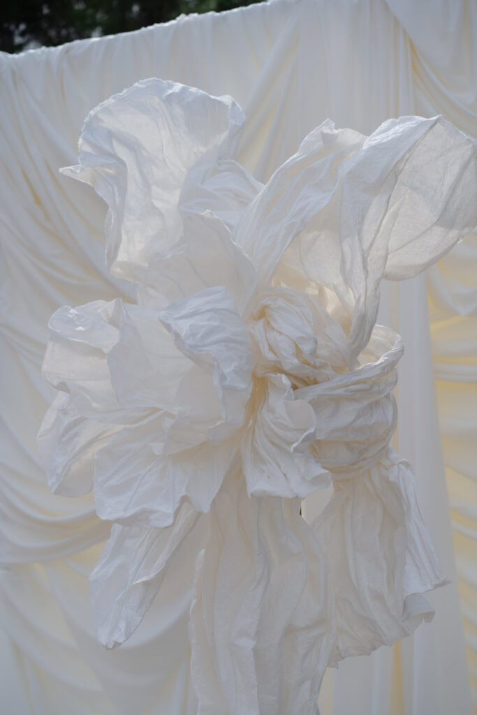 Eine große weiße Stoffblumenskulptur vor einem drapierten Stoffhintergrund, sanft beleuchtet von natürlichem Tageslicht, die künstlerisches Textildesign und kreative visuelle Elemente präsentiert.