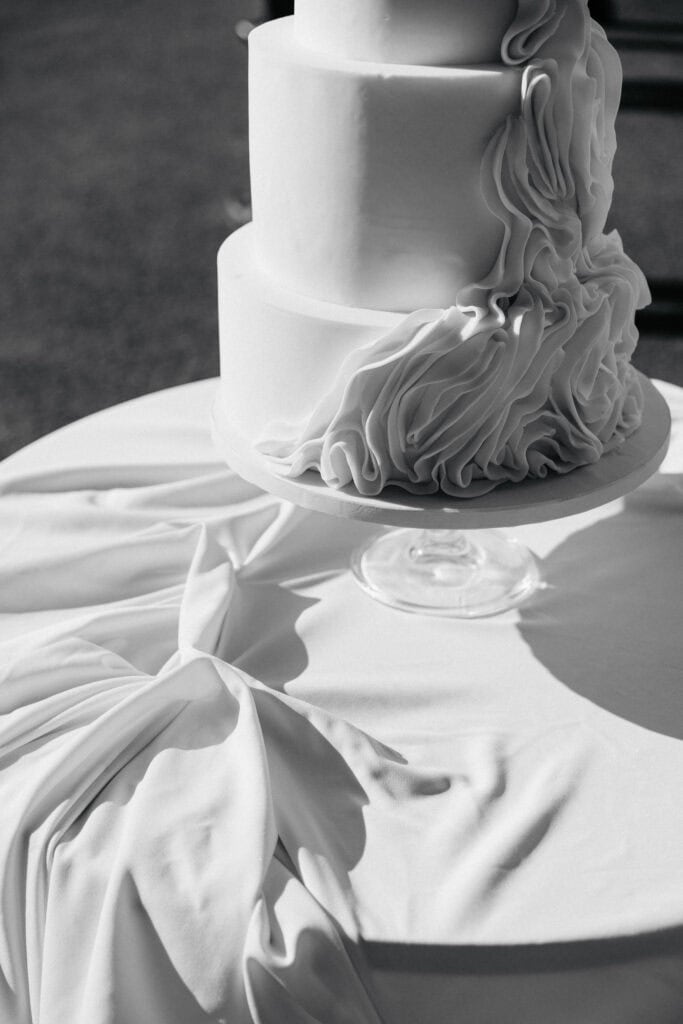 Elegante Hochzeitstorte mit gerafftem Zuckerguss und glatten Fondantschichten auf einem Glasständer, aufgenommen in Schwarz und Weiß.