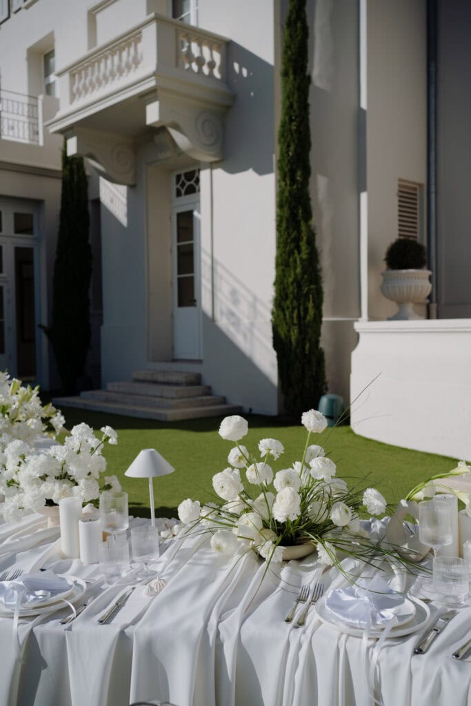 Eleganter Hochzeitstisch im Freien mit weißem Blumenschmuck und edlem Geschirr vor einem luxuriösen weißen Villenhintergrund.