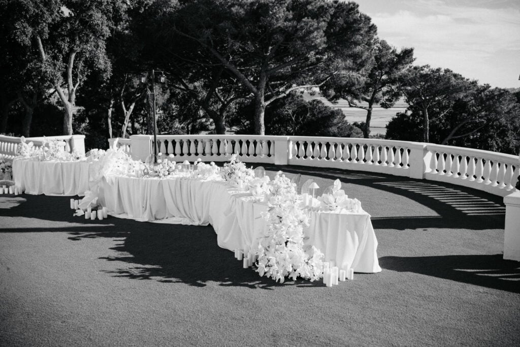Eleganter Hochzeitsempfang im Freien mit einem weiß drapierten Tisch, Blumenarrangements und Kerzenakzenten auf einer malerischen Terrasse mit Bäumen im Hintergrund.