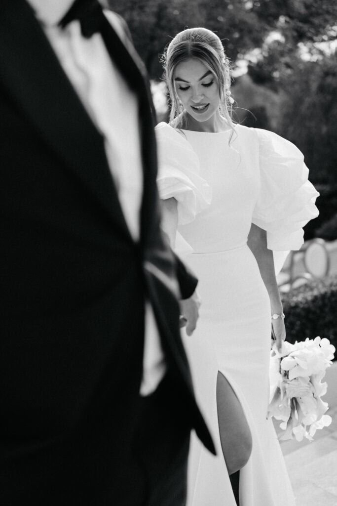 Eine Braut in einem eleganten weißen Kleid mit voluminösen Puffärmeln, die einen Blumenstrauß in der Hand hält, blickt erfreut auf ihre Hochzeitszeremonie im Freien in Castel Bay.