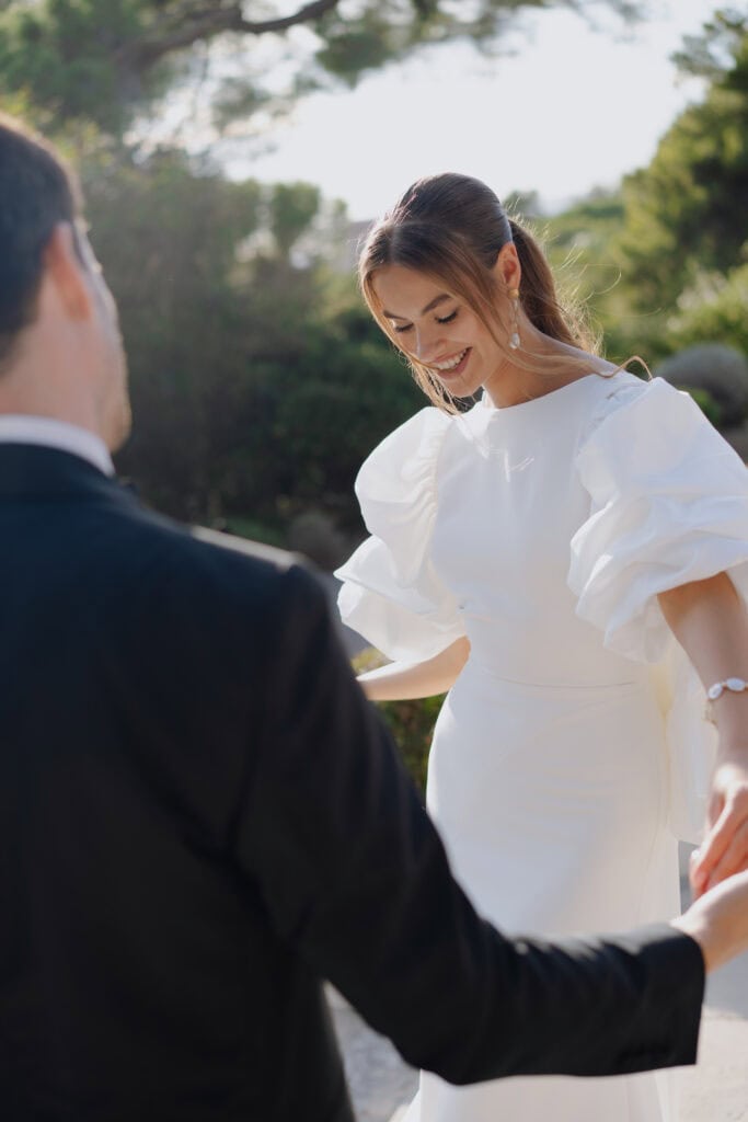 Elegante Braut in einem weißen Hochzeitskleid mit Puffärmeln, lächelt freudig während des Gelübdes im Freien mit ihrem Partner an einem sonnigen Tag in Castel Bay.
