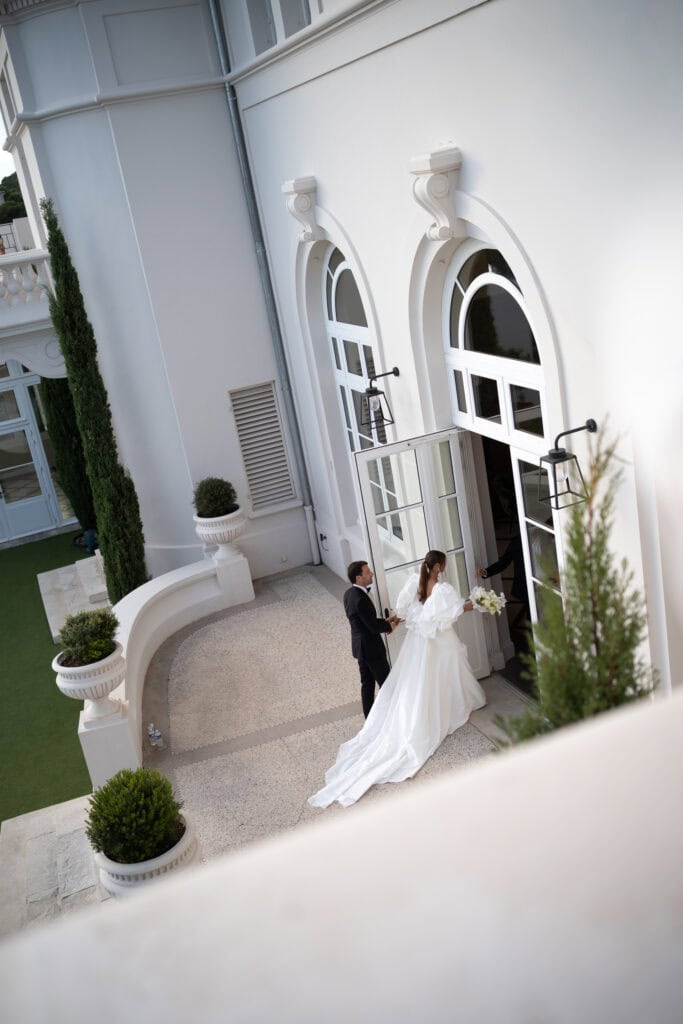 Elegante Braut im weißen Hochzeitskleid beim Betreten eines großen weißen Gebäudes mit Bogenfenstern und klassischem Dekor.