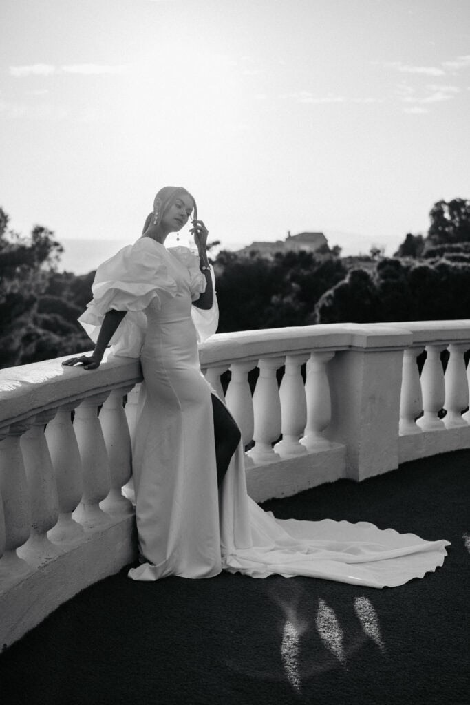 Elegante Frau in einem langen weißen Kleid auf einem Dachbalkon, mit raffinierter Mode und malerischer Aussicht, ideal für stilvolle Bilder von Hochzeiten oder Abendveranstaltungen.