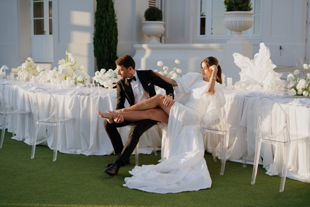 Elegantes Hochzeitspaar auf Stühlen sitzend bei einer Feier im Freien mit weißem Blumendekor und drapierten Tischtüchern.