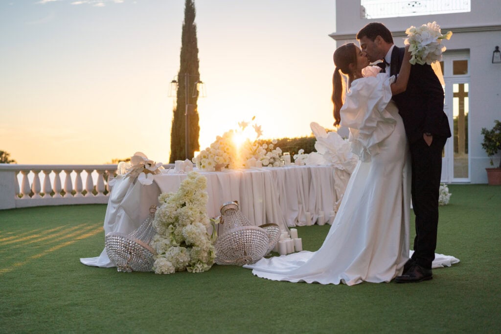 Ein frisch verheiratetes Paar küsst sich romantisch während seiner Hochzeitsfeier im Freien bei Sonnenuntergang, wobei elegante Blumenarrangements und Dekoration den besonderen Moment unterstreichen.