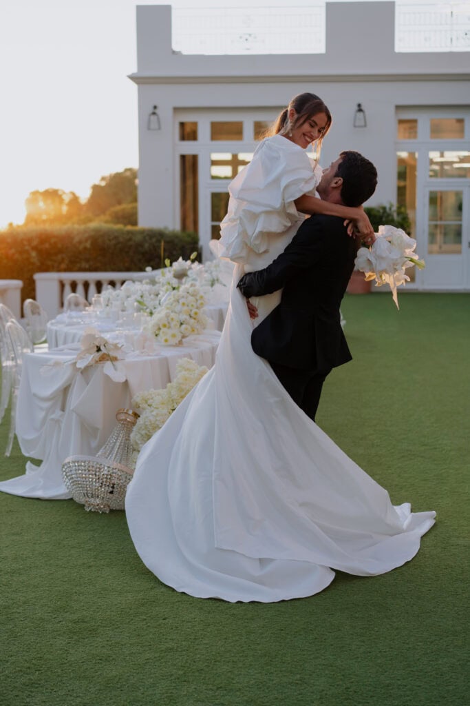 Ein fröhliches Paar feiert seine Hochzeit im Freien bei Sonnenuntergang, mit eleganten Tischen und Blumendekorationen im Hintergrund. Die Braut trägt ein wunderschönes weißes Kleid, während der Bräutigam sie in einer romantischen Pose hochhebt.