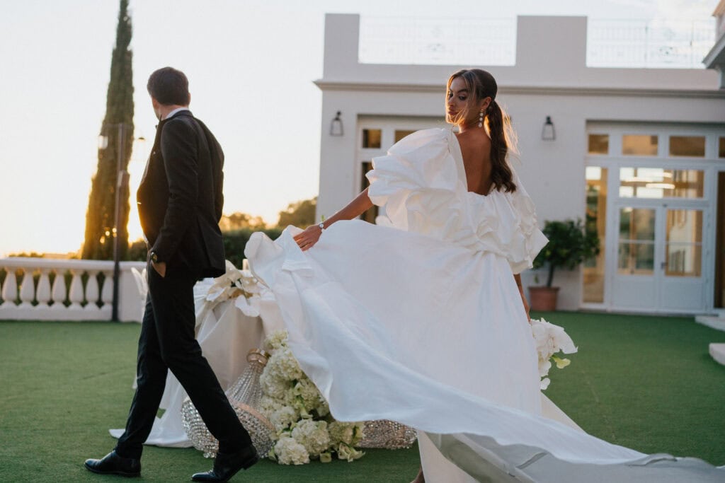 Elegante Braut in einem dramatischen weißen Hochzeitskleid mit Puffärmeln und fließender Schleppe, stehend auf einem üppigen grünen Rasen während der goldenen Stunde im Freien Hochzeit.