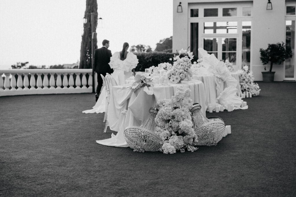 Eleganter Hochzeitsempfang im Freien mit dekorierten Tischen, Blumenschmuck und luxuriösen Details auf einem üppigen Rasen. Perfekt für eine anspruchsvolle Feier oder Verlobungsfeier. Hochwertige Veranstaltungsdekoration mit raffinierten Blumenarrangements und stilvollen Akzenten.