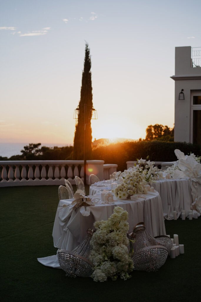 Eleganter Hochzeitsempfang im Freien mit weißen Blumenarrangements bei Sonnenuntergang und luxuriöser Veranstaltungsdekoration auf einer malerischen Terrasse mit Meerblick.
